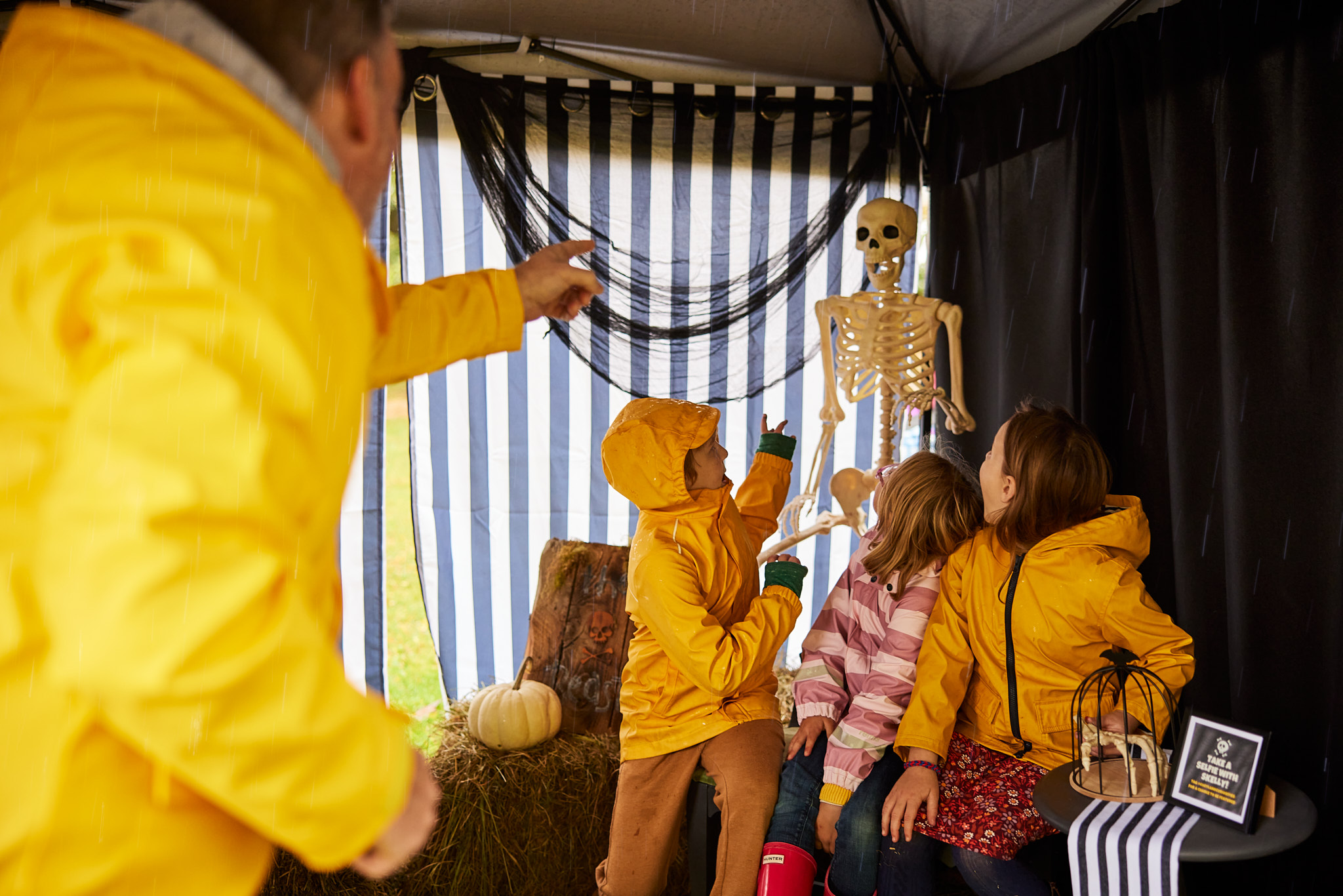 family in yellow rain coats posing with large, fake skeleton for photos under a black and white tent and Cortland Pumpkinfest.